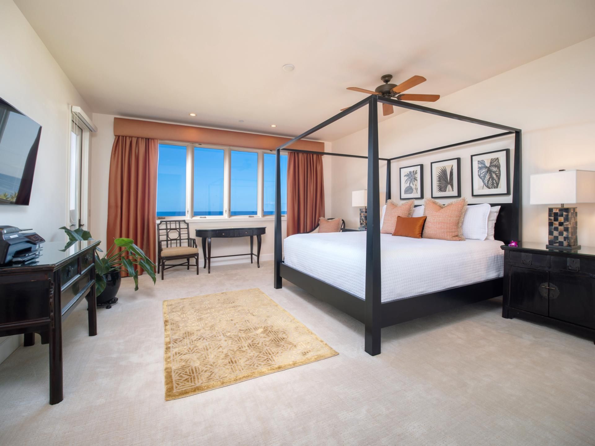 A four-poster bed with a ceiling fan, a desk with a chair, a view of the ocean, and framed artwork on the wall.