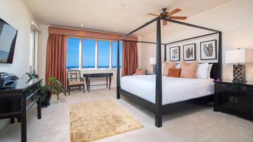 A four-poster bed with a ceiling fan, a desk with a chair, a view of the ocean, and framed artwork on the wall.