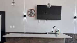 Una cocina moderna con una isla grande, taburetes de bar, luces colgantes, un reloj en la pared y un televisor de pantalla plana.