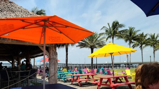 A beachside dining area with colorful tables and umbrellas, overlooking the ocean. - Ideal location - 2bed/1bath near the beach & shops (Port Charlotte)