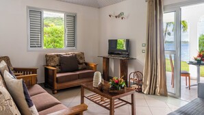 A cozy living room with a sofa, a coffee table with a decorative item, a television on a stand, and a view of a lush garden through a window. - Paradise (Bora Bora)