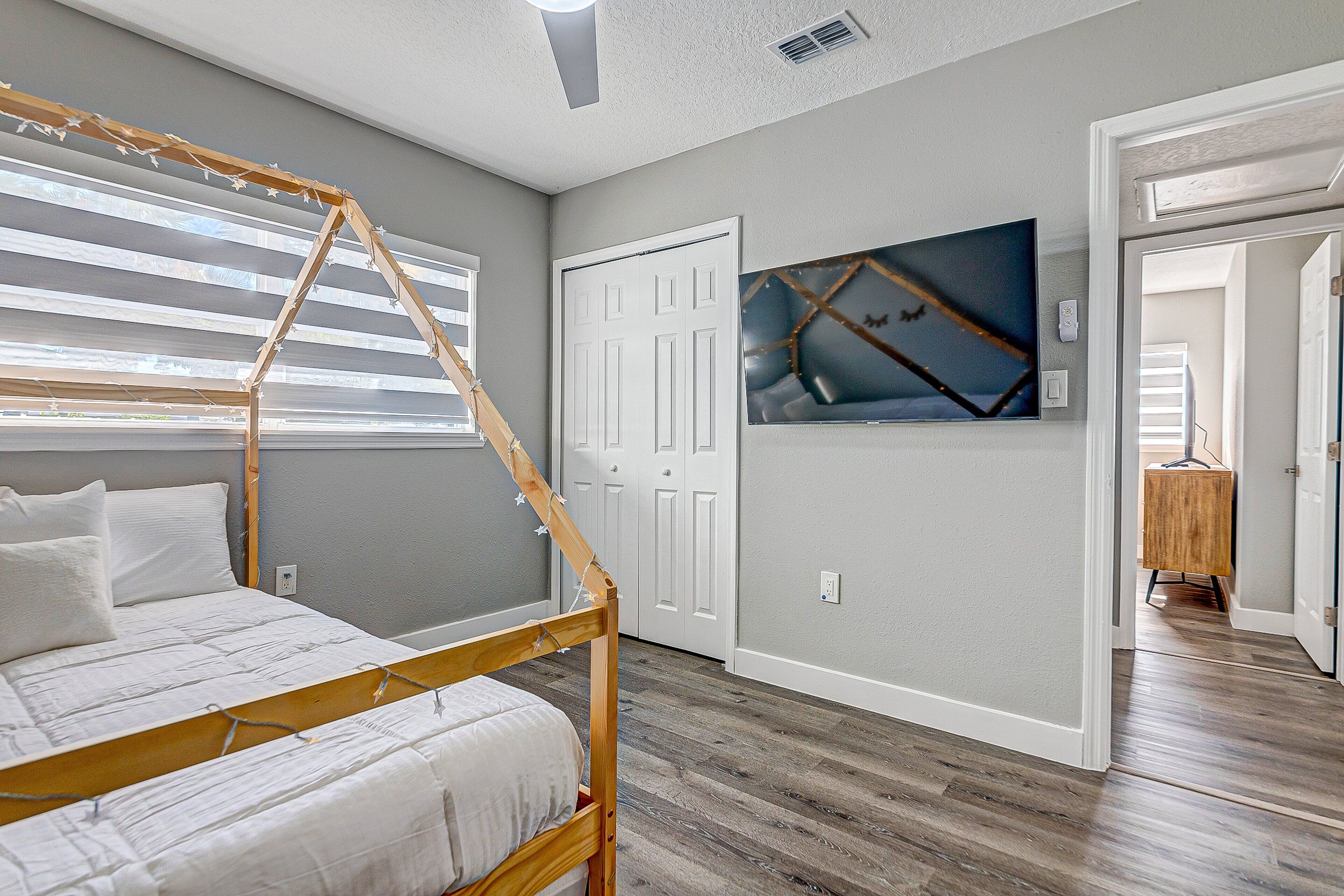 A bedroom with a wooden four-poster bed, a large window with blinds, a door, and a wall-mounted artwork.