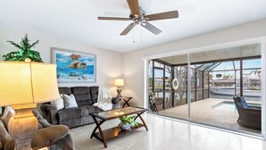Ein modernes Wohnzimmer mit Deckenventilator, einer Couch, einem Couchtisch und einem großen Fenster mit Blick auf einen Poolbereich.