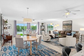 A spacious living room with a dining area, a ceiling fan, and a large window offering a view of greenery. - Cocoa Beach Riverfront Retreat — Screened Porch, Wildlife Views & Short Drive to the Beach! 🌅 (Cocoa Beach)