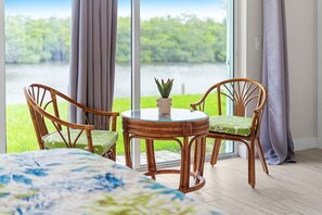 A room with a bed, a round table with chairs, a potted plant, and a view of a lake through large windows. - Cocoa Beach Riverfront Retreat — Screened Porch, Wildlife Views & Short Drive to the Beach! 🌅 (Cocoa Beach)