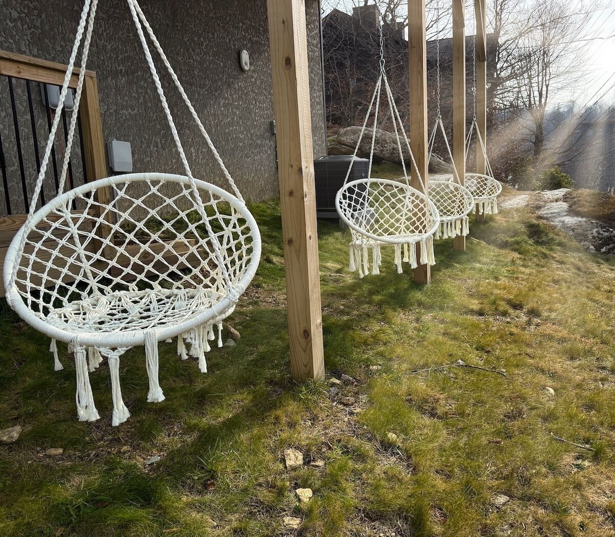 Three white macrame hanging chairs suspended from wooden posts in a grassy outdoor area.