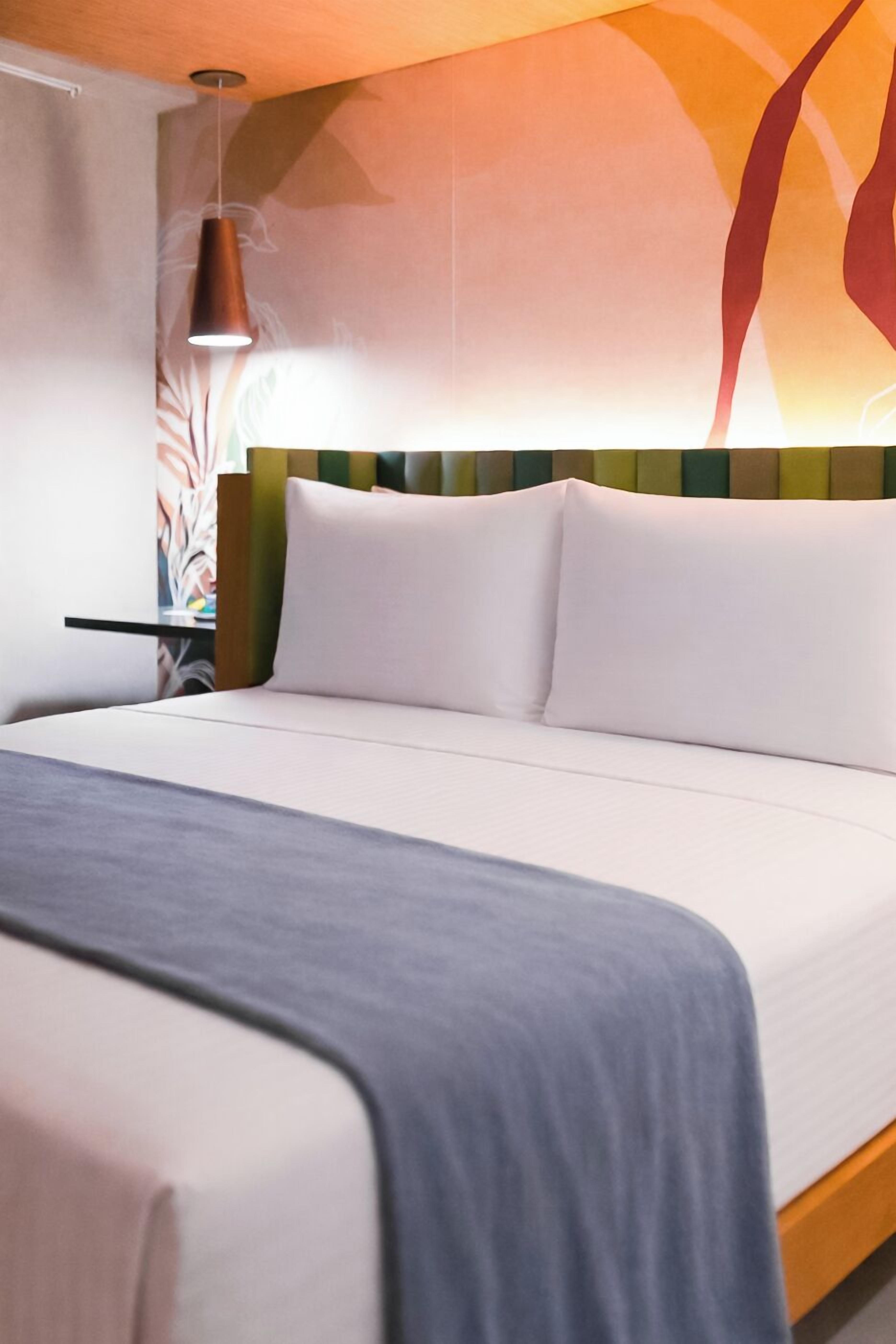 Una habitación de hotel moderna con una cama, una mesita de noche, una lámpara y una pared decorada con arte abstracto.