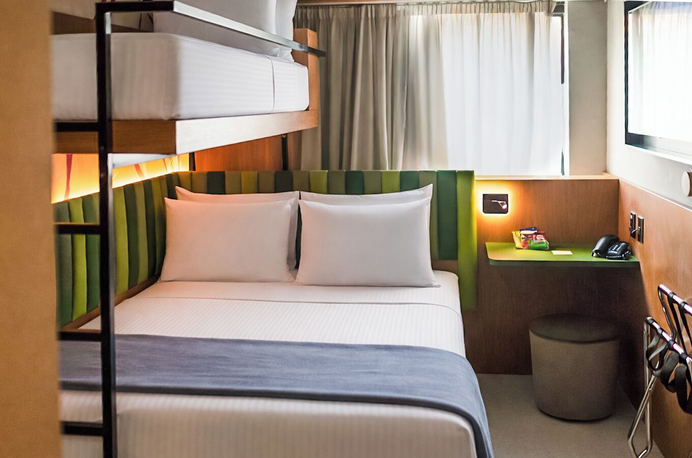 Una cama nido con cabecera verde, una mesita de noche con un teléfono y una pequeña caja, un cesto de basura y una ventana con cortinas.