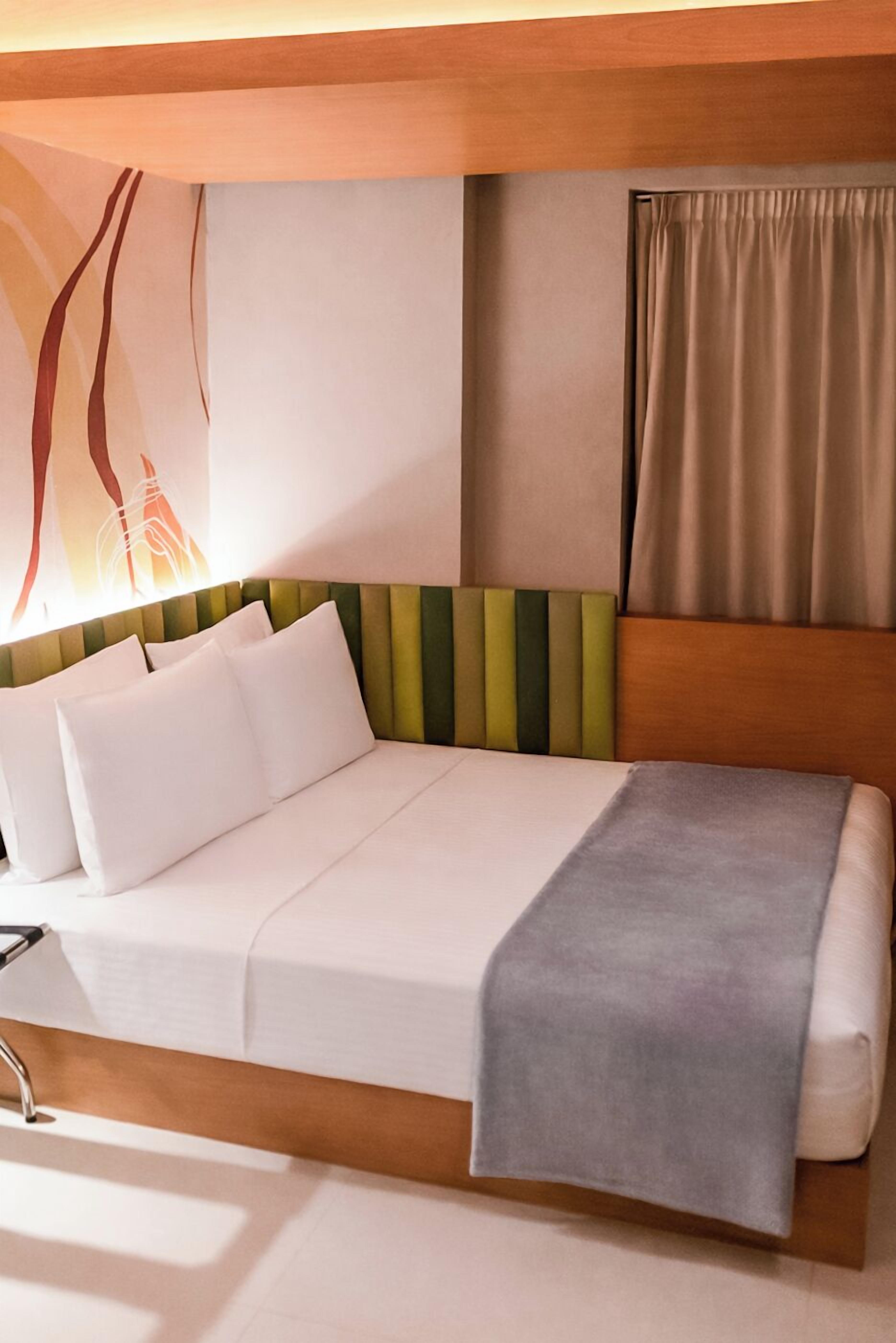 Quarto de hotel moderno com cama, cabeceira com listras verticais, mesa de cabeceira e luminária fixada na parede.