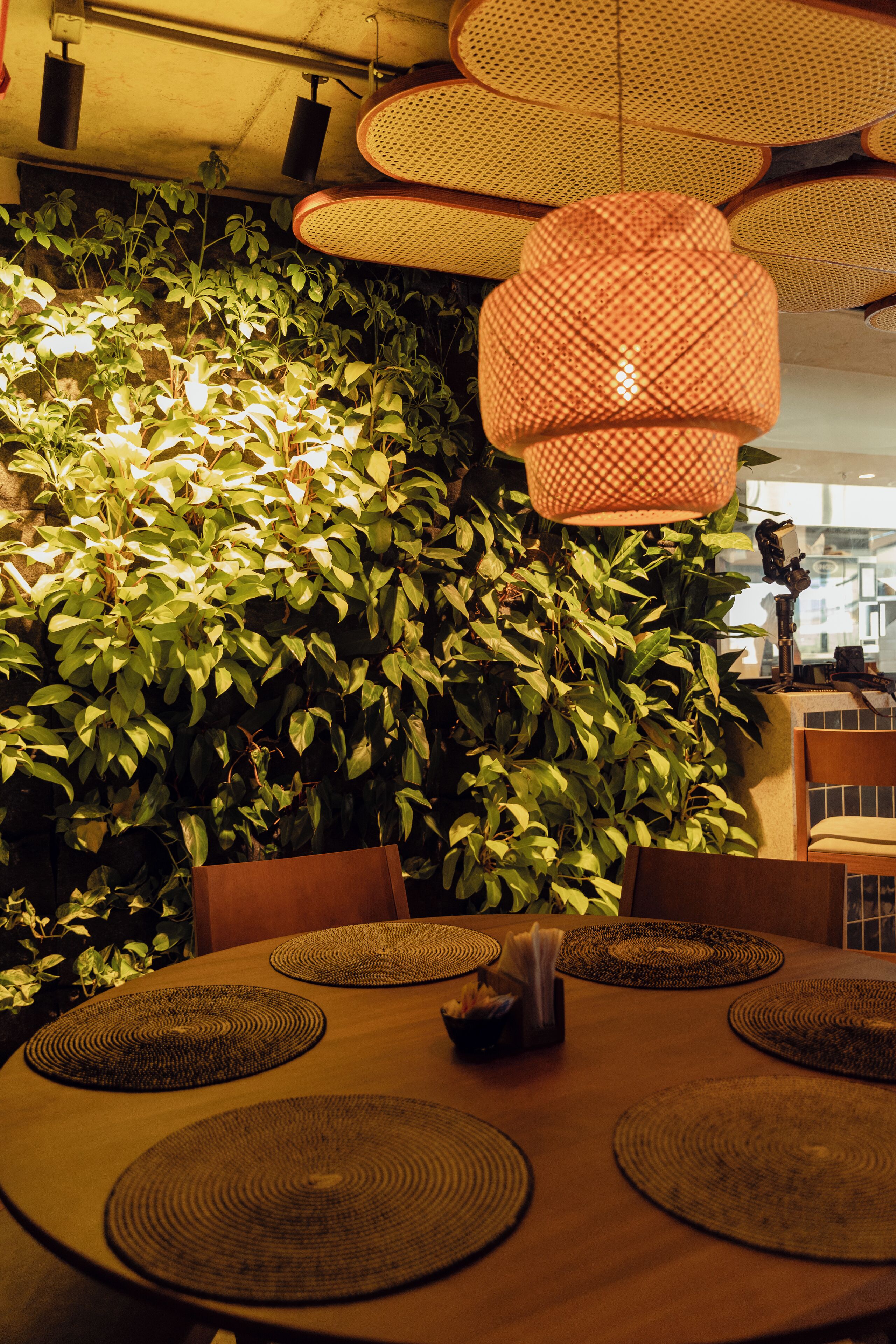 A dining area with a round table, woven placemats, and a wall of green foliage.