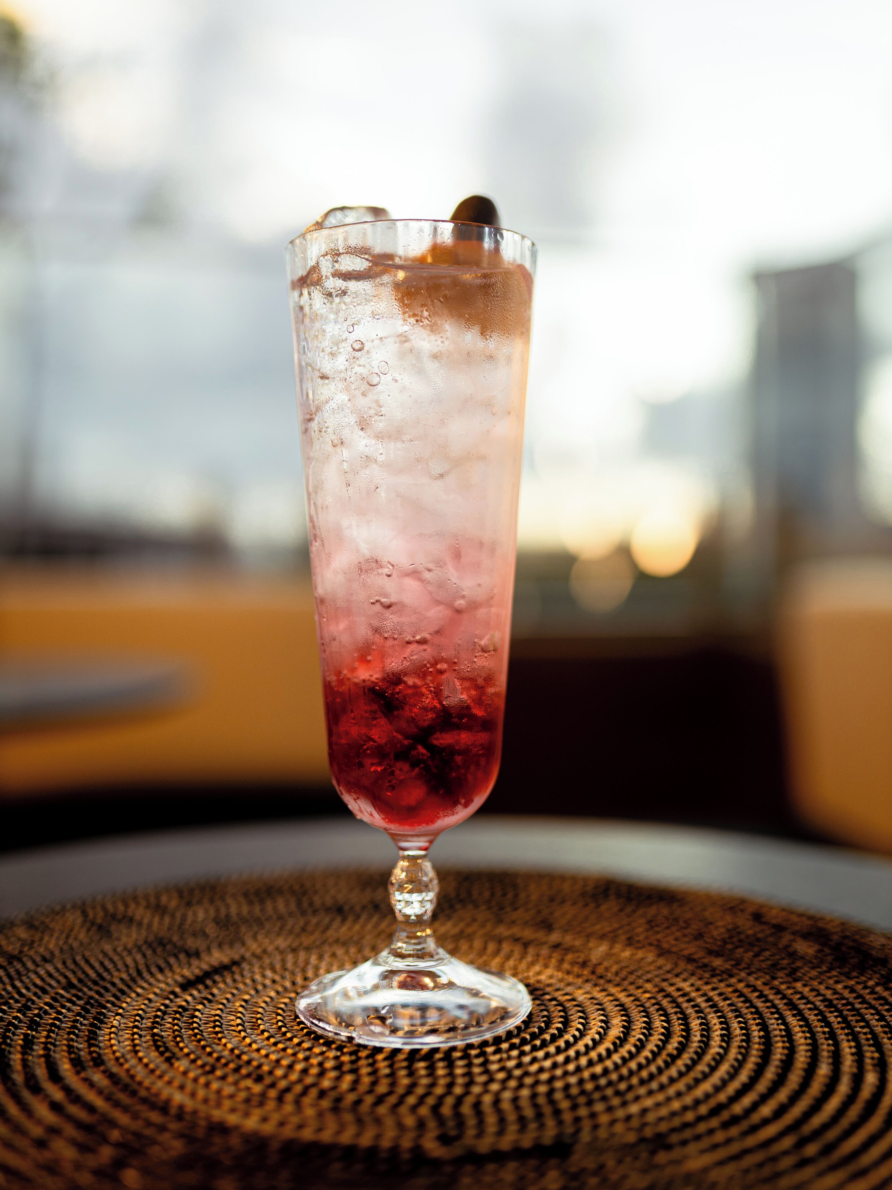 A tall glass with a red drink and ice, placed on a woven mat.