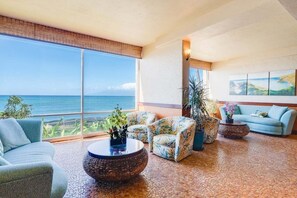 A spacious living area with a large window overlooking the ocean, a blue sofa, patterned armchairs, and a round coffee table. - Spectacular ocean views at Spacious 2-BR AC Pool- Royal Kahana 610 (Lahaina)
