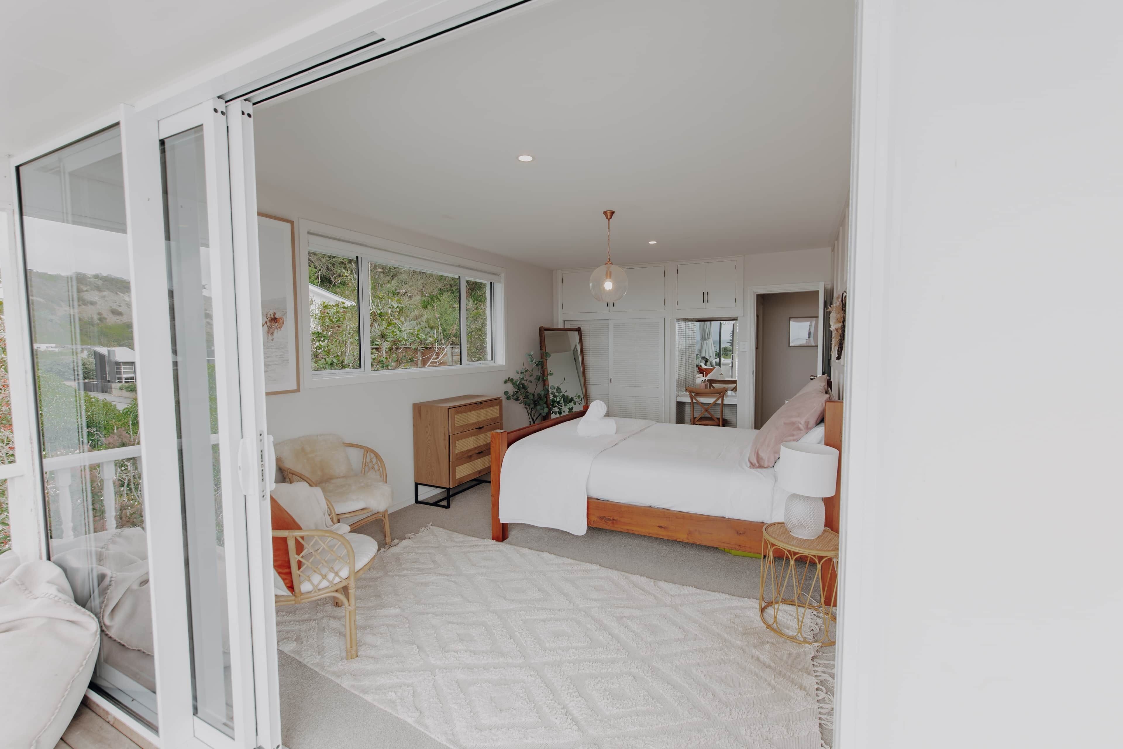 A modern bedroom with a large bed, a wooden dresser, a chair, and a glass door leading to a balcony.