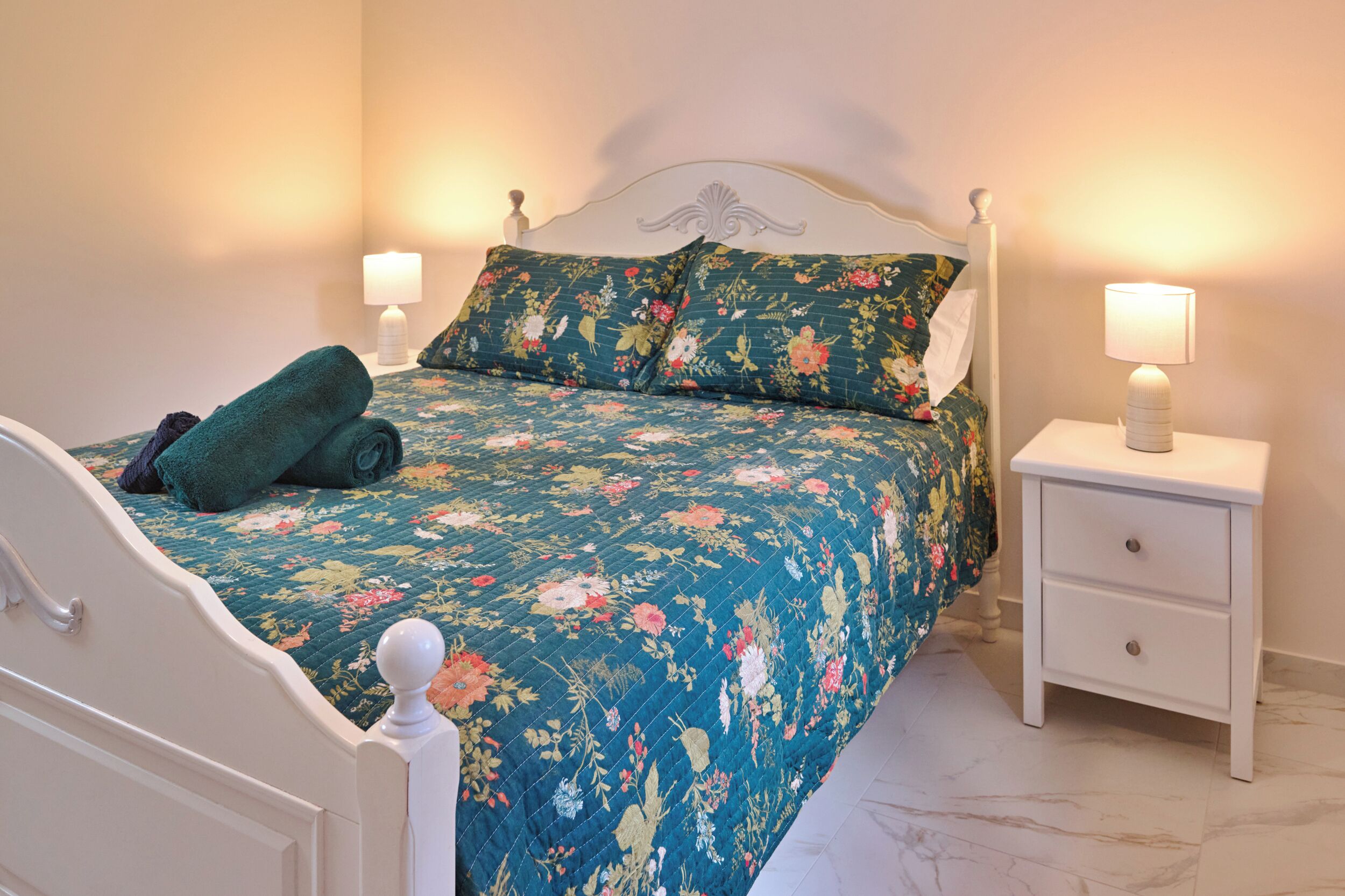 A bed with a floral bedspread, two pillows, a rolled-up towel, and a bedside table with a lamp.