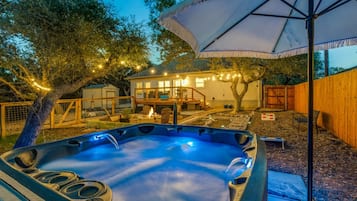 Uma banheira de hidromassagem com um deck de madeira e um guarda-sol, rodeada por árvores e uma casa com iluminação externa.