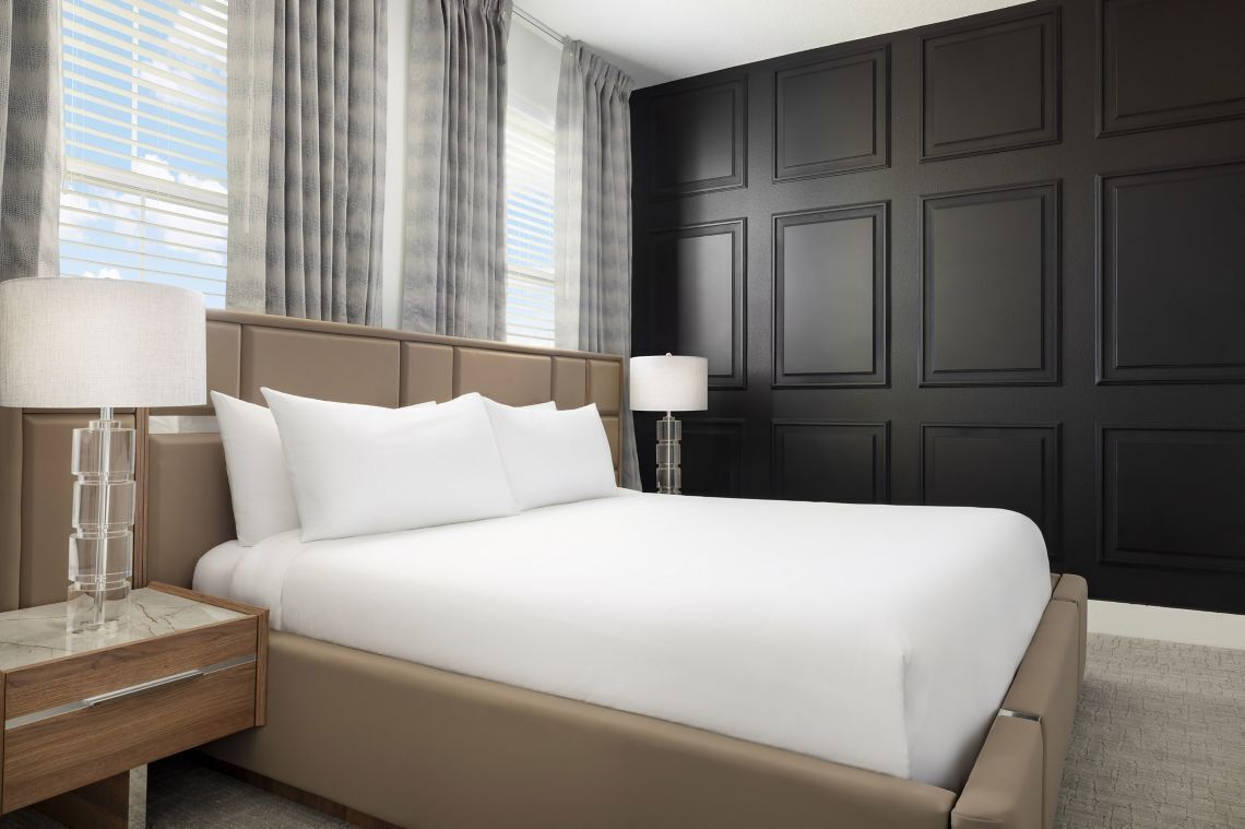 A hotel room with a large bed, two bedside tables with lamps, a dark paneled wall, and a window with curtains.