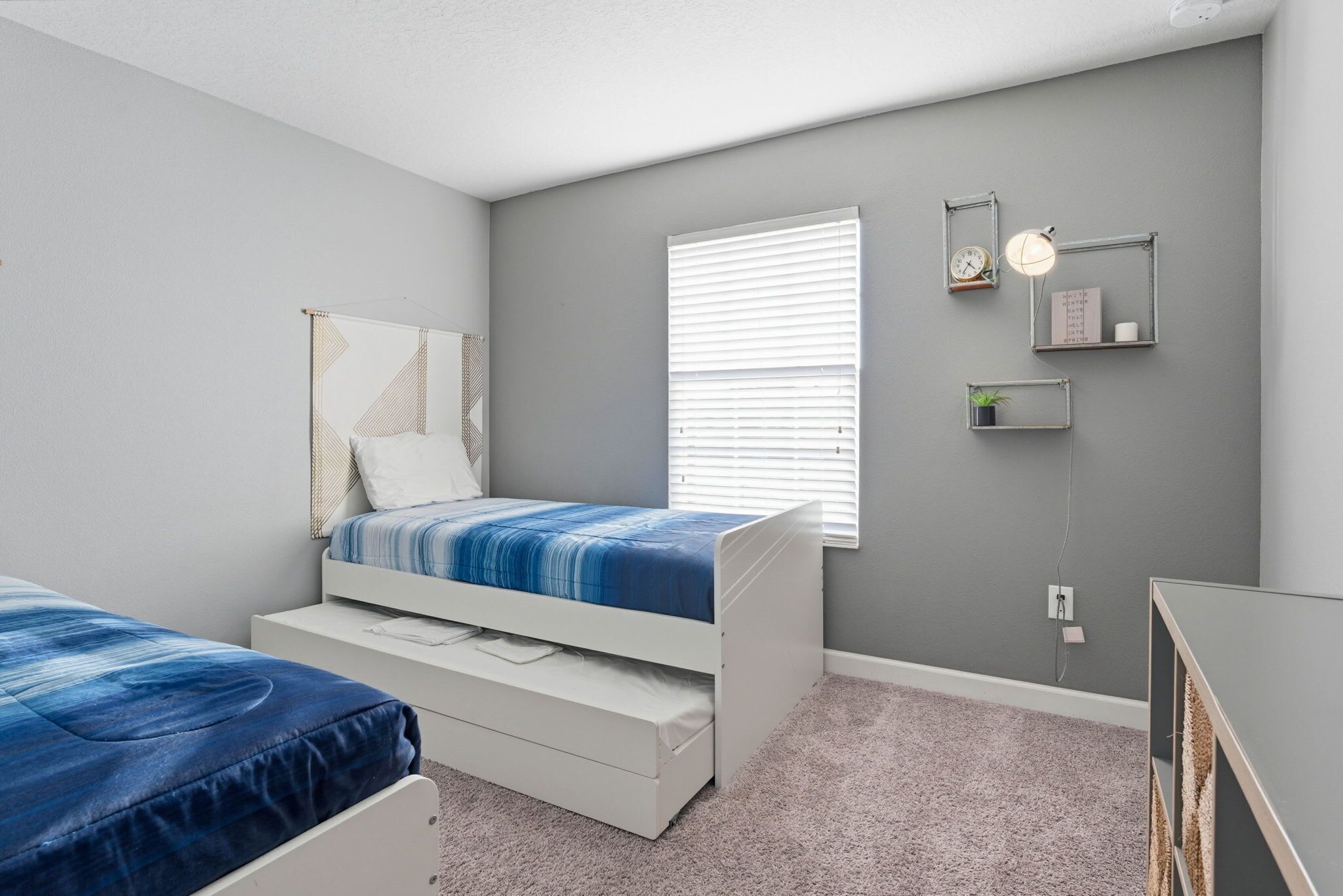A bedroom with two beds, a window with blinds, a bedside table, a mirror, and wall-mounted shelves with clocks and a plant.
