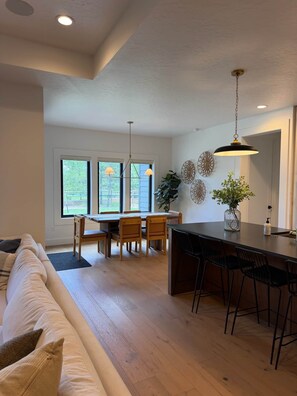 A spacious living area with a sofa, dining table, and chairs, a kitchen island with bar stools, and large windows offering a view of the outdoors. - Cabin w/Hot Tub & Bunk Room - Steps to the lake! (Valley County)