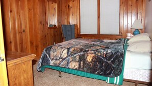 A bedroom with a wooden paneled wall, a bed with a camouflage bedspread, a chair, a nightstand with a lamp, and a window with blinds. - Delightful 3-bedroom cabin on Big Roche Cri Lake in charming Friendship, WI! (Friendship)