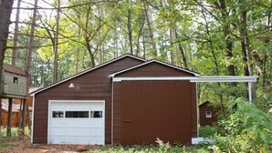 A brown single-story building with a white garage door, surrounded by trees. - Delightful 3-bedroom cabin on Big Roche Cri Lake in charming Friendship, WI! (Friendship)