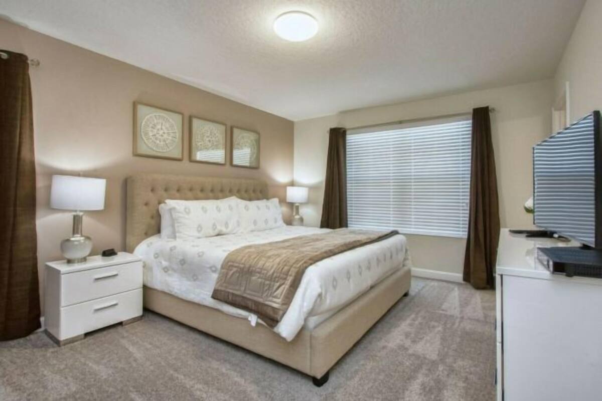 A bedroom with a large bed, a nightstand, a desk, and a window with curtains.