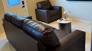 Un salón con un sofá de cuero marrón, un sillón marrón, una mesita redonda pequeña, un cuadro en la pared y un televisor montado sobre el sillón.