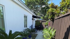 Un sendero de concreto estrecho bordeado por plantas en macetas y una valla de madera, que conduce a una casa de fachada blanca y un porche cubierto.