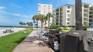 A wooden deck with a grill and seating area, overlooking a beach and a hotel building. - 🌊 New Listing! Caprice Resort Unit #305 – Gulf Front Comfort with Private Balcony (Saint Pete Beach)