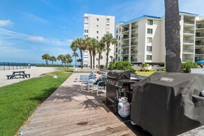 A wooden deck with a grill and seating area, overlooking a beach and a hotel building. - 🌊 New Listing! Caprice Resort Unit #305 – Gulf Front Comfort with Private Balcony (Saint Pete Beach)