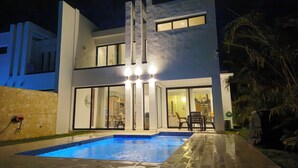 Une maison moderne à deux étages, dotée d’une piscine, de grandes fenêtres et d’une entrée bien éclairée.
