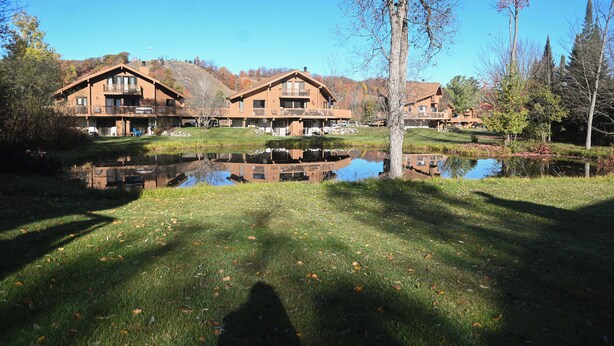 A large, two-story lodge with a balcony on the second floor, a pond in front, and a clear blue sky. - New rental! Schuss. Walk to slopes sleeps 4, walk to slopes, Renovated kitchen,  (Mancelona)