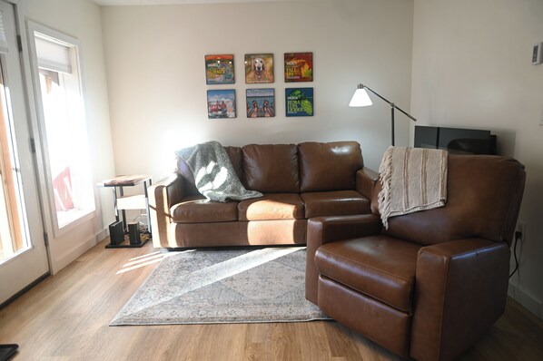 Een woonkamer met een bruine leren bank, een bijzettafeltje, een lamp en een vloerkleed. Er hangen zes ingelijste foto's aan de muur.