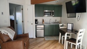 A compact kitchen with green cabinets, a microwave, and a refrigerator. There is a dining table with chairs and a TV mounted on the wall. - New rental! Schuss. Walk to slopes sleeps 4, walk to slopes, Renovated kitchen,  (Mancelona)
