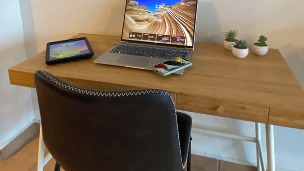 A wooden desk with a laptop, a smartphone, and a tablet. There are three small potted plants on the desk.