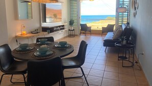 A modern living room with a dining area, a large TV, and a view of the beach.