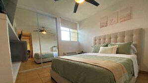 Una camera da letto con un letto grande, una ventola a soffitto, una TV e una porta scorrevole che dà su un'altra stanza.