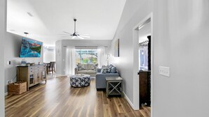 Uma sala de estar moderna com piso de madeira, um sofá cinza, um banquinho azul com estampas de estrelas, uma TV de tela plana e um ventilador de teto.