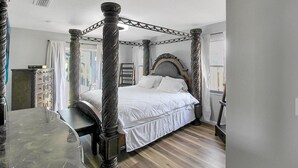 Uma cama de dossel com colcha branca, um banquinho de madeira e uma mesa de mármore.