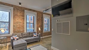 A living room with a grey sectional sofa, a small side table, and a lamp. There are large windows with views of buildings outside. - Sleek Studio Near Dtwn Dining & Entertainment (Cincinnati)