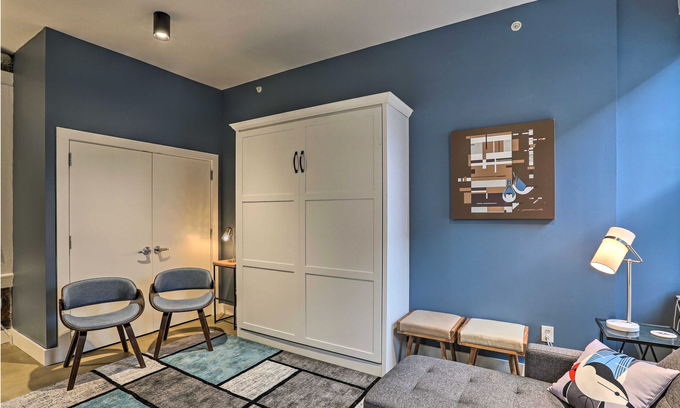 Een kamer met een blauwe wand, een witte kast, twee blauwe stoelen, een klein tafeltje met een lamp en een bank met kussens.