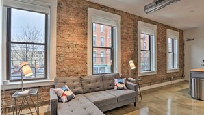 A spacious room with a grey sofa, two floor lamps, and large windows with a brick wall. - Sleek Studio Near Dtwn Dining & Entertainment (Cincinnati)