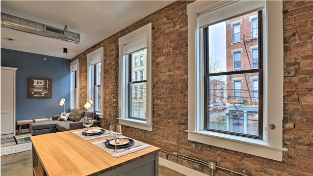 A modern kitchen with a wooden table set for dining, large windows with a view of the city, and exposed brick walls. - Sleek Studio Near Dtwn Dining & Entertainment (Cincinnati)