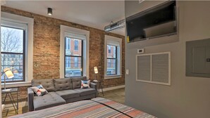 A modern living room with a brick wall, large windows, a grey sectional sofa, and a striped area rug. - Sleek Studio Near Dtwn Dining & Entertainment (Cincinnati)