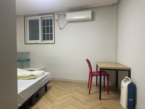 A small, sparsely furnished room with a bed, a desk, a chair, and an air conditioning unit mounted on the wall.