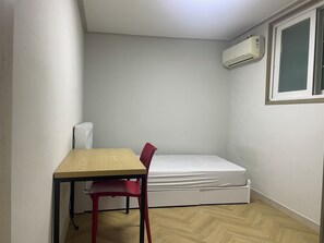 A small, sparsely furnished room with a wooden table, a red chair, a white mattress, and a window with an air conditioning unit.