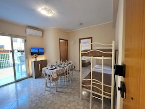 Une chambre d’hôtel avec un lit superposé, un coin repas avec une table dressée pour deux personnes, une télévision et une porte-fenêtre coulissante donnant sur l’extérieur.