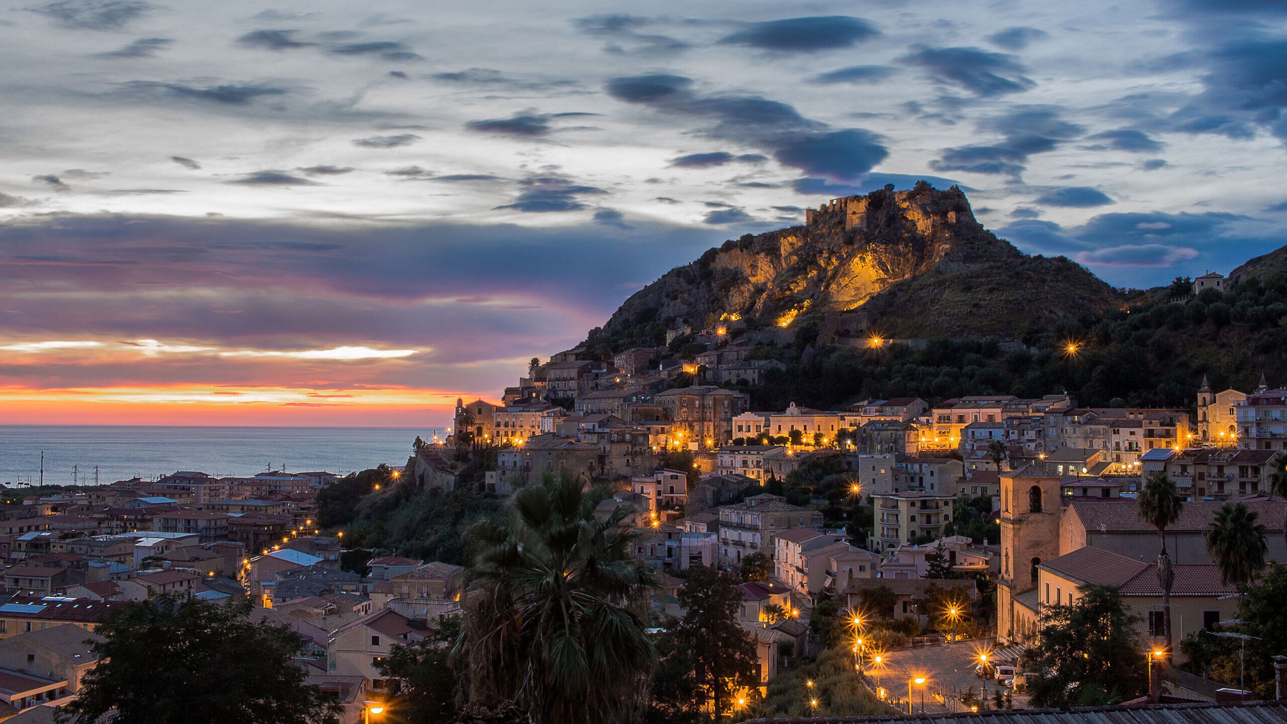 Une ville côtière au crépuscule, avec des bâtiments historiques, une structure imposante située sur une colline et une vue sur la mer.