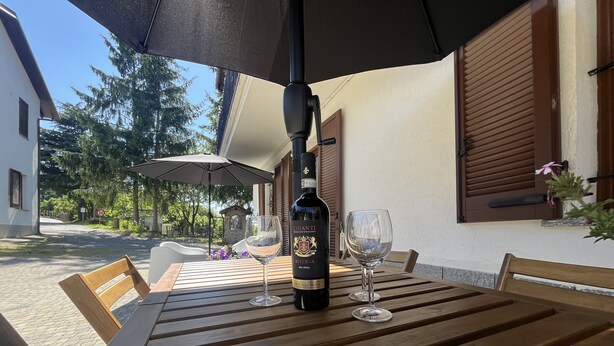 A wooden table with a bottle of wine and two glasses, under an umbrella. - Panoramic Estate in the Langhe with Pool - Up to 12 Guests, Ideal for Groups (Piemonte)