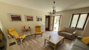 A living room with wooden flooring, a sofa, two yellow armchairs, a coffee table, and a TV stand. There are three framed pictures on the wall, a chandelier, and a window with curtains. - Panoramic Estate in the Langhe with Pool - Up to 12 Guests, Ideal for Groups (Piemonte)