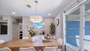 A modern dining area with a wooden table, white chairs, a neon sign, and a large window. - Welcoming 3-bedroom house in sunny Naples: Beach Days Made Easy (Naples)